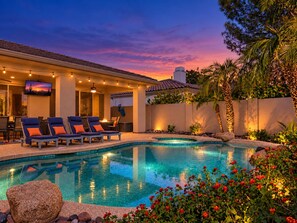 A swimming pool with lounge chairs and a television screen, surrounded by a landscaped garden with palm trees and flowering plants.