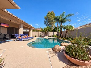 A pool area with lounge chairs, a television, and a landscaped garden with palm trees and rocks.
