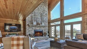 A cozy living room with a stone fireplace, large windows, and a flat-screen TV. - Carter`s Lake Overlook | Ellijay, Ga (Ellijay)