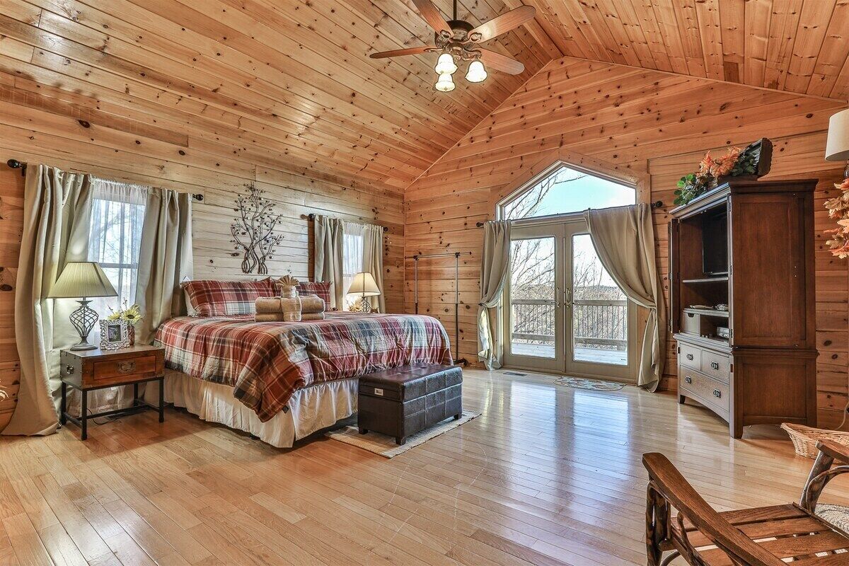 A log cabin bedroom with a bed, bedside lamps, a dresser, and a large window.