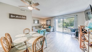 A spacious living room with a dining area, a ceiling fan, and a large window offering a view of the outdoors.
