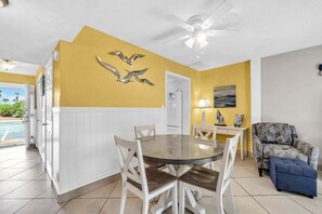 A dining area with a round glass table and white chairs, a ceiling fan, and a yellow wall with a bird sculpture.