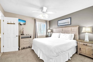 A bedroom with a large bed, a ceiling fan, a window with curtains, a bedside table with a lamp, and a door with a coat rack.