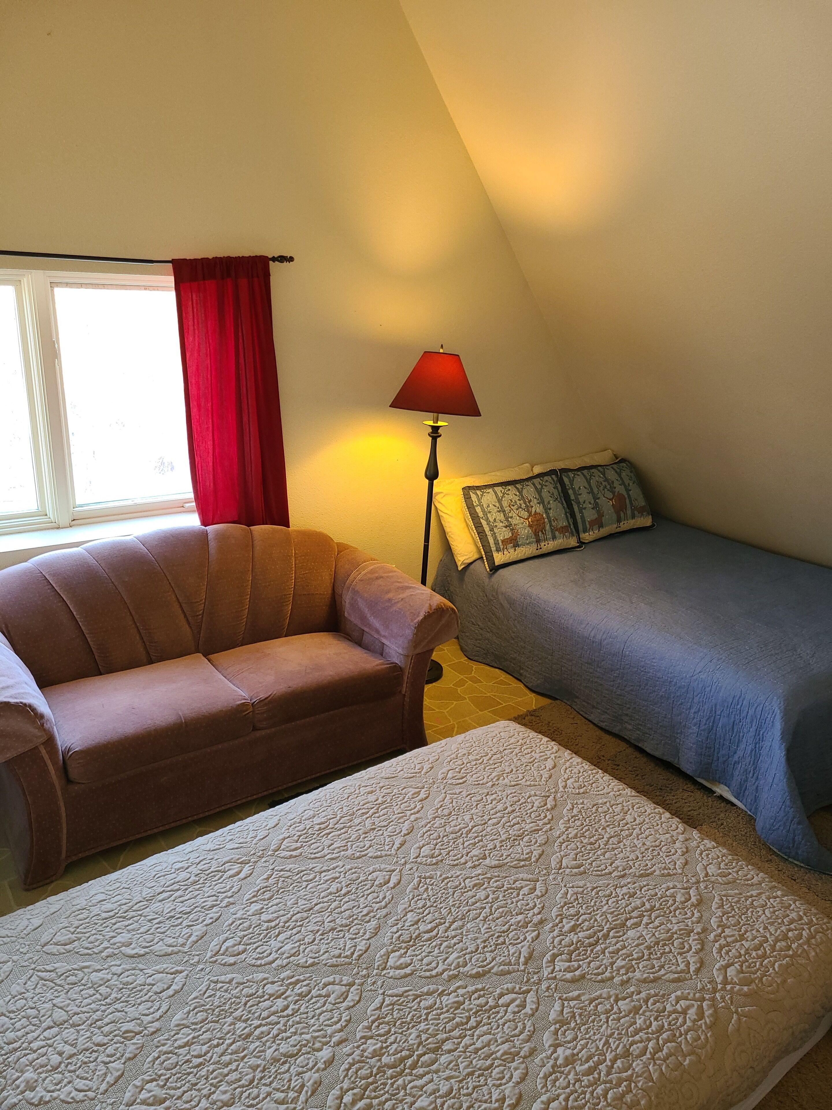 Une pièce chaleureuse avec un canapé marron, un petit lit recouvert d’une couverture bleue, une lampe avec un abat-jour rouge et une fenêtre ornée de rideaux rouges.