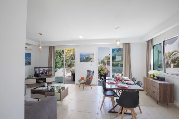 A modern living room with a TV, seating, a dining area, and large windows with a view.
