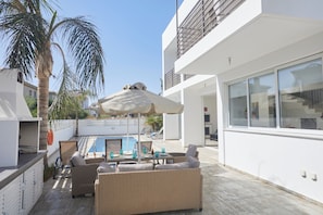 A modern outdoor patio with wicker furniture, a pool, and a white building with large windows. - Protaras Pearl Villa Pe17 (Paralimni)