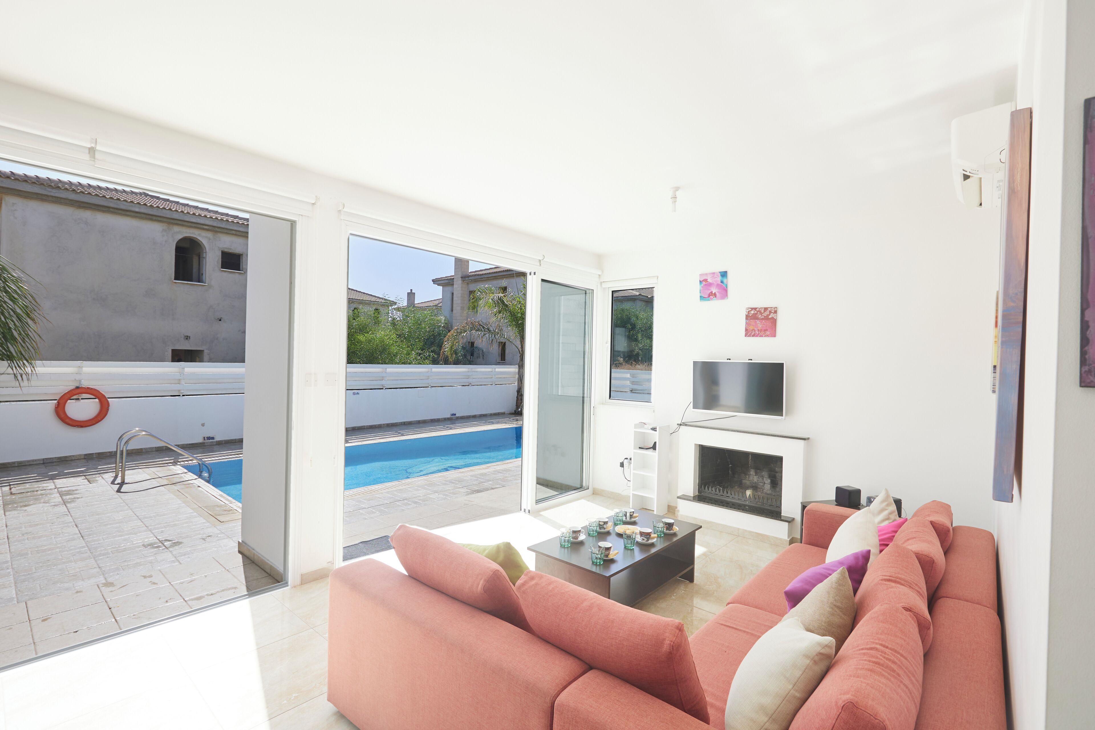 A modern living room with a sofa, a coffee table, and a fireplace. There is a pool visible through the glass doors.