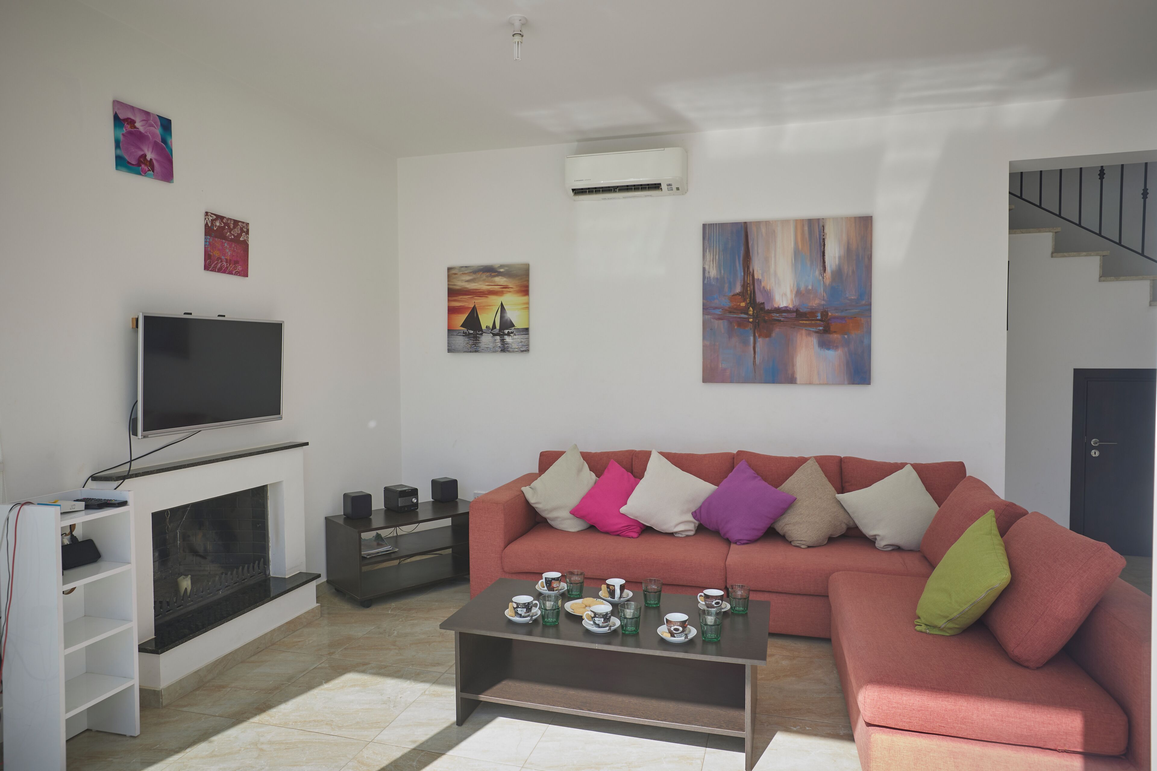 Ein modernes Wohnzimmer mit Flachbildfernseher, Kamin, einer Couch mit bunten Kissen und einem Couchtisch mit Tassen und Untertellern.
