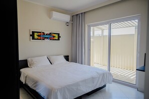 A bedroom with a bed, a wall-mounted air conditioner, a sliding glass door to a balcony, and a piece of abstract art.