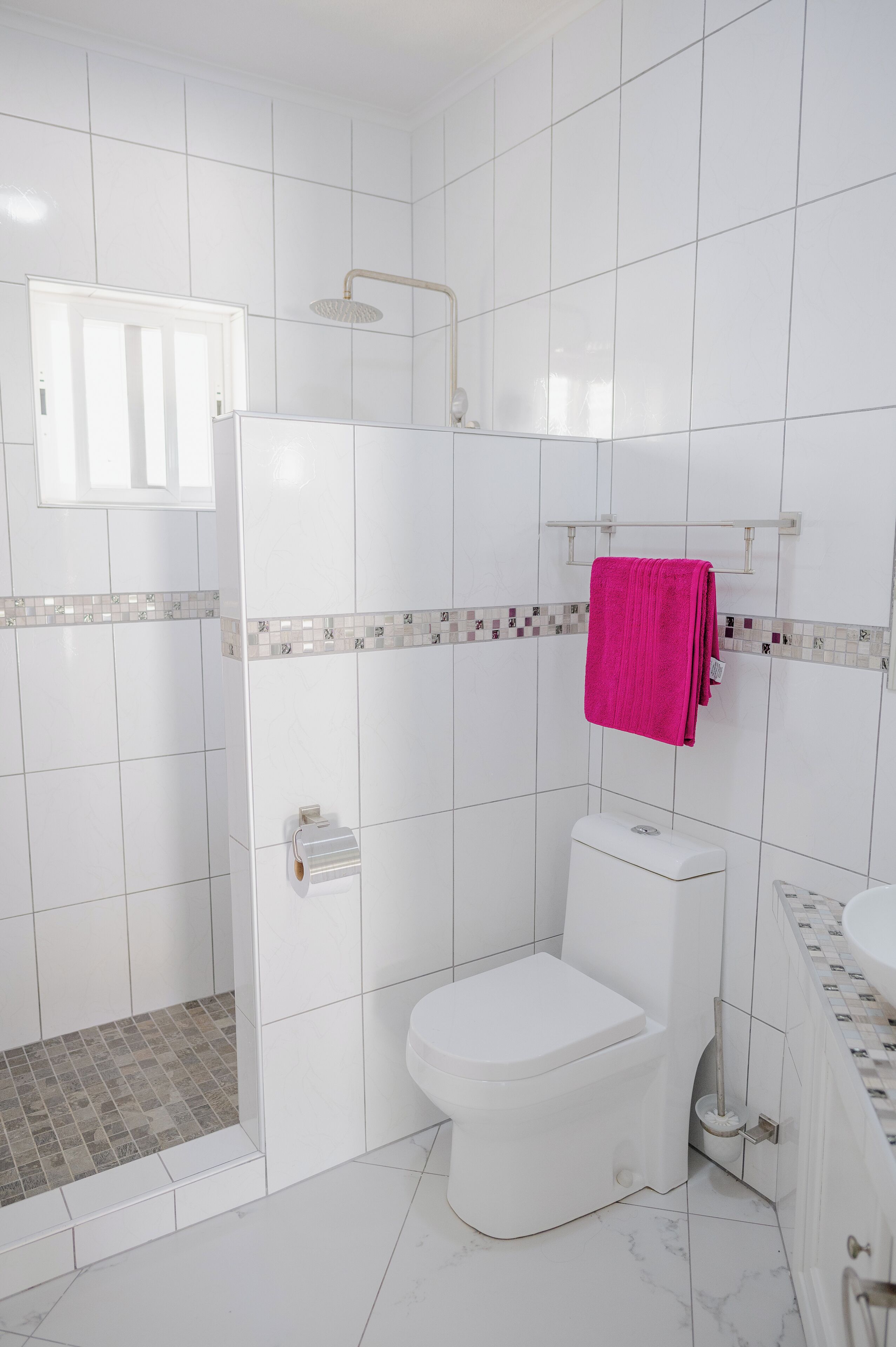 Un baño con azulejos blancos, inodoro blanco, ducha con puerta de cristal y una toalla rosa colgada en la pared.