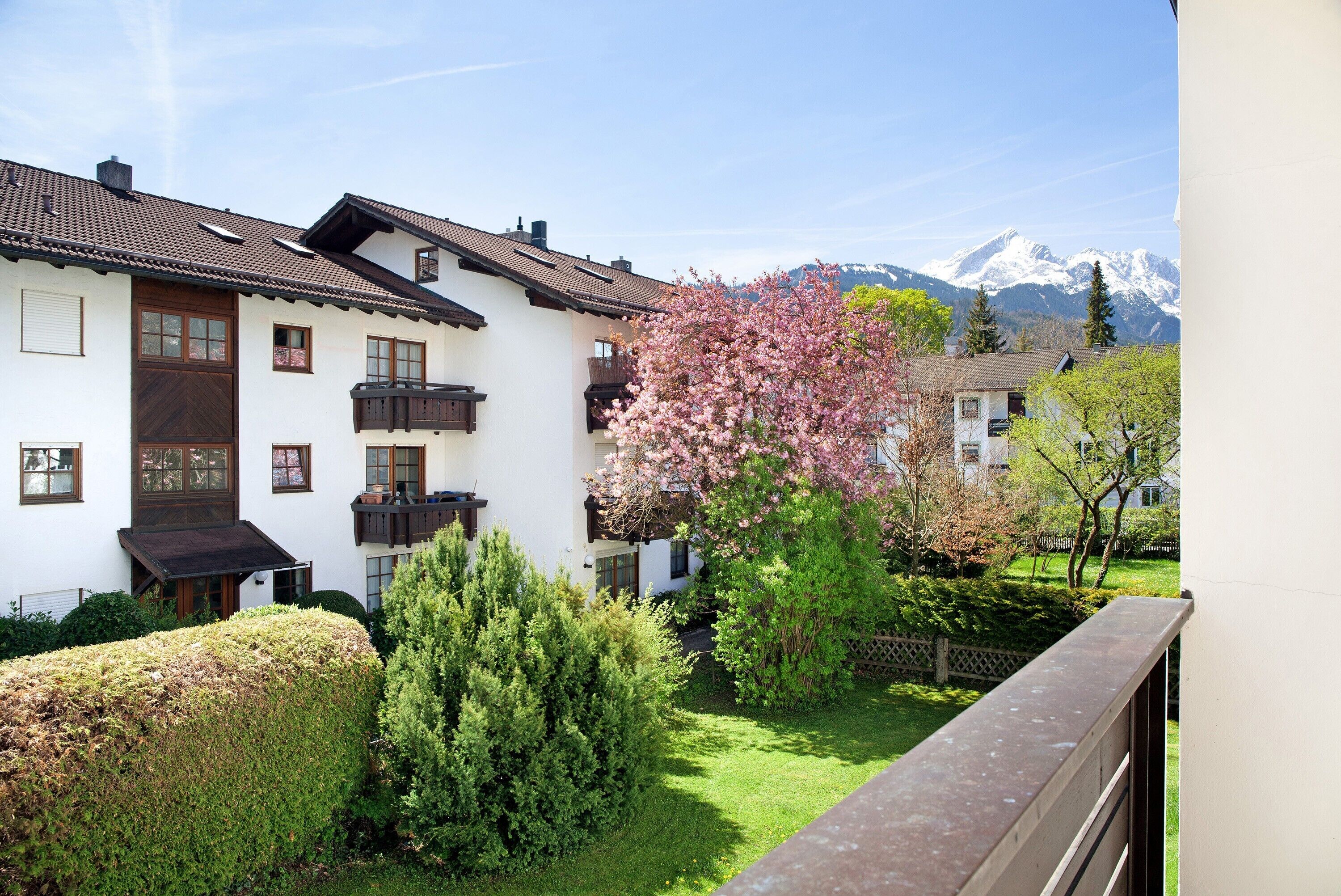 A white building with a brown roof, a balcony, and a garden with a pink flowering tree.