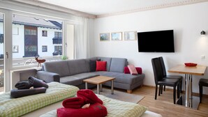 Una sala moderna con un sofá gris, una mesa de centro de madera, un televisor montado en la pared y un balcón con vistas a un edificio.