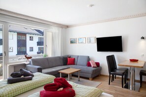 Una sala moderna con un sofá gris, una mesa de centro de madera, un televisor montado en la pared y un balcón con vistas a un edificio.