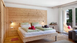 Un dormitorio con cabecera de madera, cama con ropa de cama blanca, una mesita de noche pequeña y vistas al exterior a través de un ventanal grande.