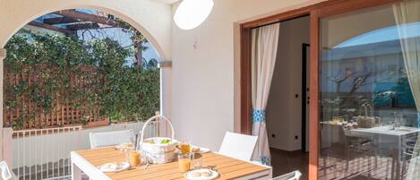 A wooden table with white chairs on an outdoor patio, a glass door leading to a balcony, and a view of a garden.
