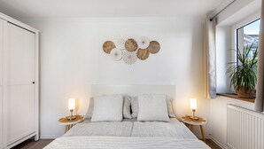 Un dormitorio con una cama grande, dos mesitas de noche con lámparas, un adorno en la pared y una ventana con una planta.