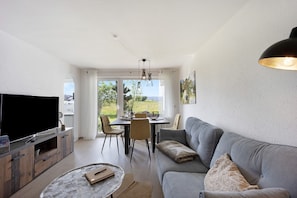 A living room with a sofa, TV, dining table, chairs, and a view through large windows. - Landliebe – Studio Apartment Near Lake with Sauna (Petersthal)
