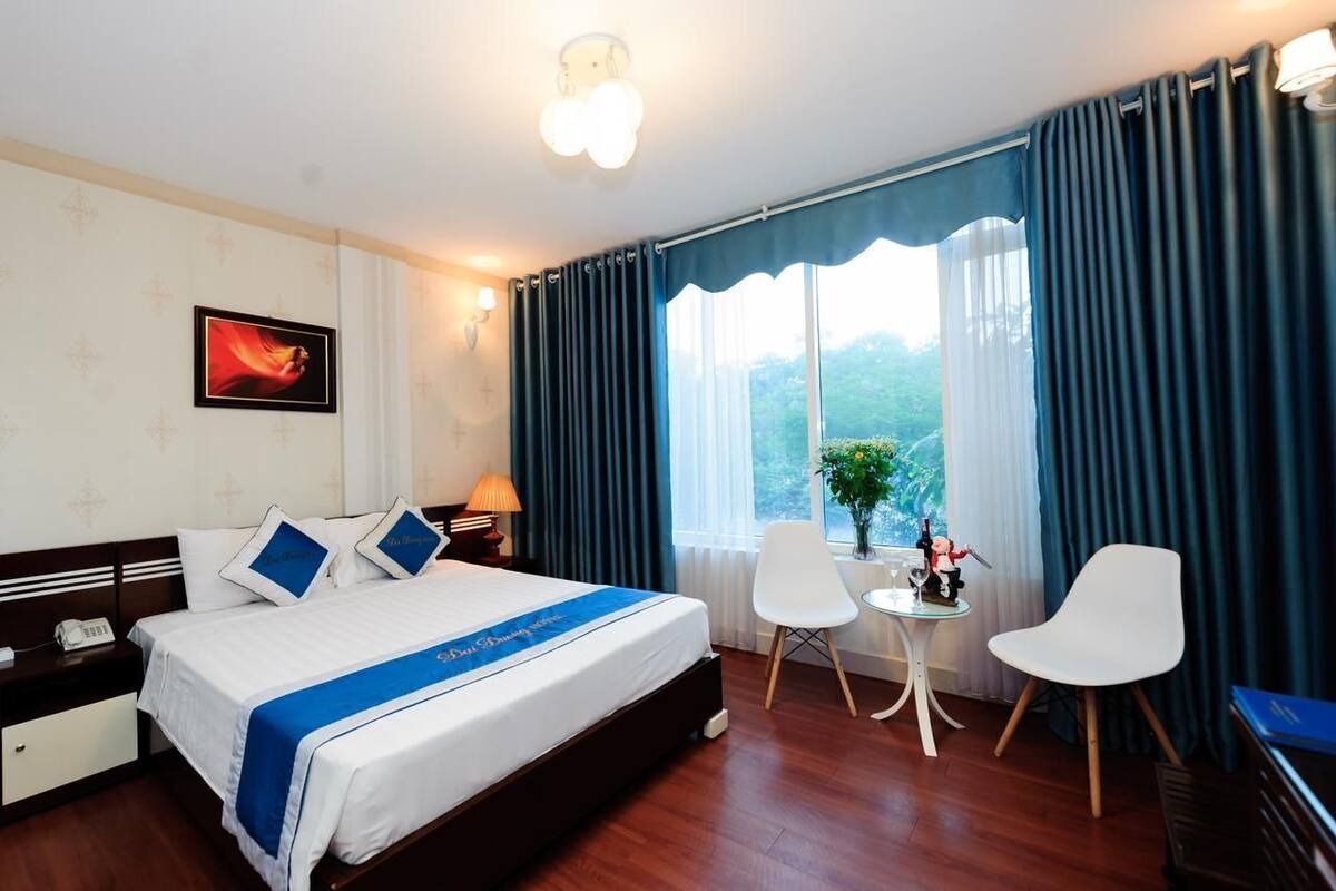 Habitación de hotel con cama, mesita de noche, lámpara, ventana con cortinas y una pequeña mesa con una silla.