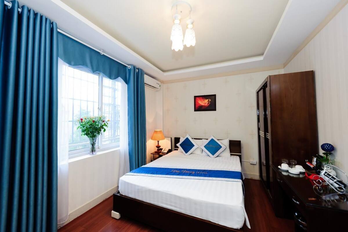 Habitación de hotel con cama, mesita de noche, lámpara, ventana con cortinas, aire acondicionado de pared, armario de madera y una pequeña mesa con un florero.