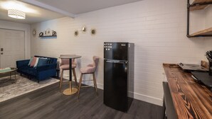 Un espacio habitable compacto con kitchenette, un sofá azul, dos sillas, una mesita y un refrigerador negro.