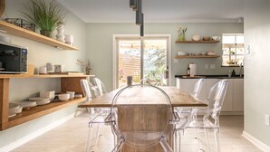 Un coin repas avec une table en bois et des chaises transparentes, une cuisine avec des étagères au-dessus du plan de travail, et une vue sur l’extérieur par une porte.