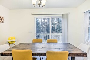 Un coin repas avec une table en bois, quatre chaises jaunes, une chaise haute jaune et une grande fenêtre donnant sur des arbres.