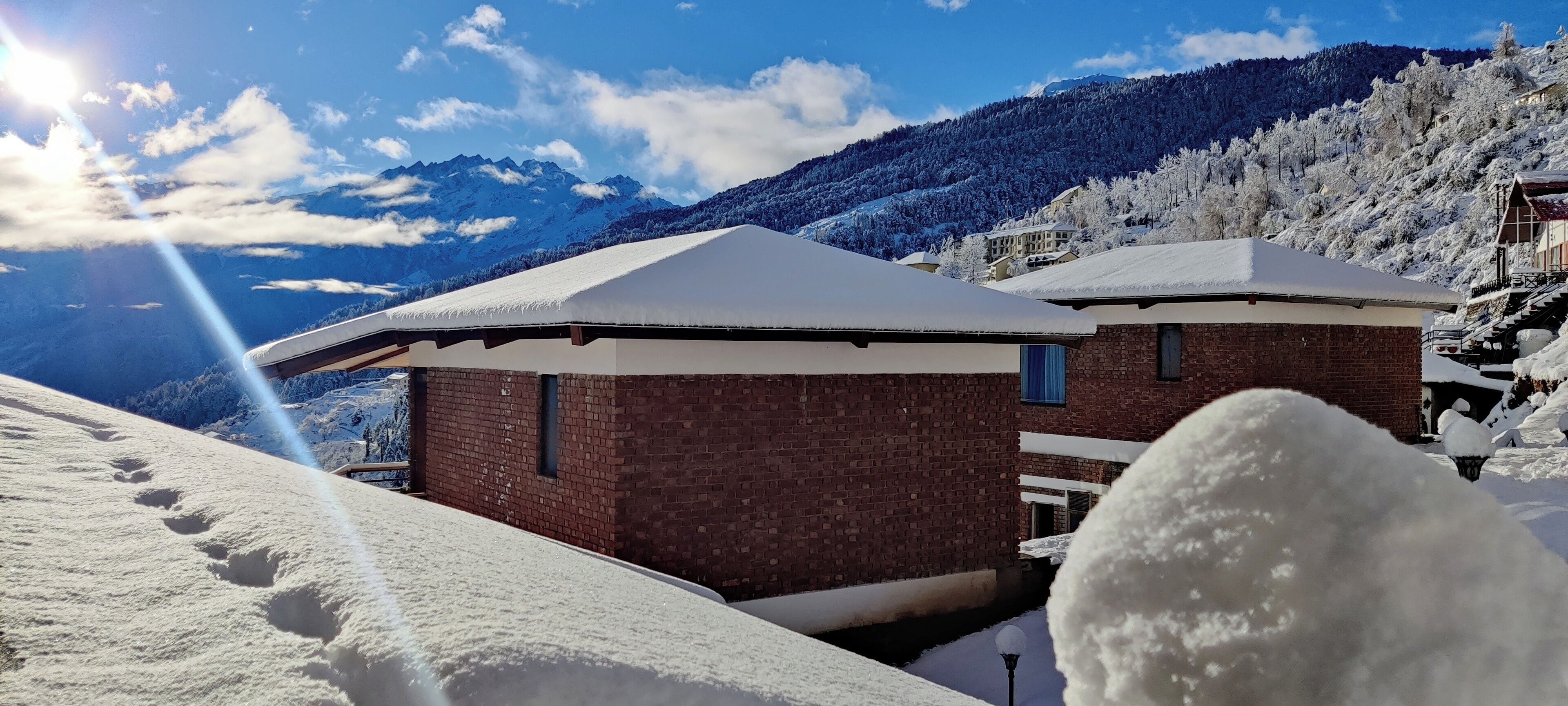 A snow-covered building with a brick facade and a flat roof, surrounded by snow-covered trees and mountains in the background.