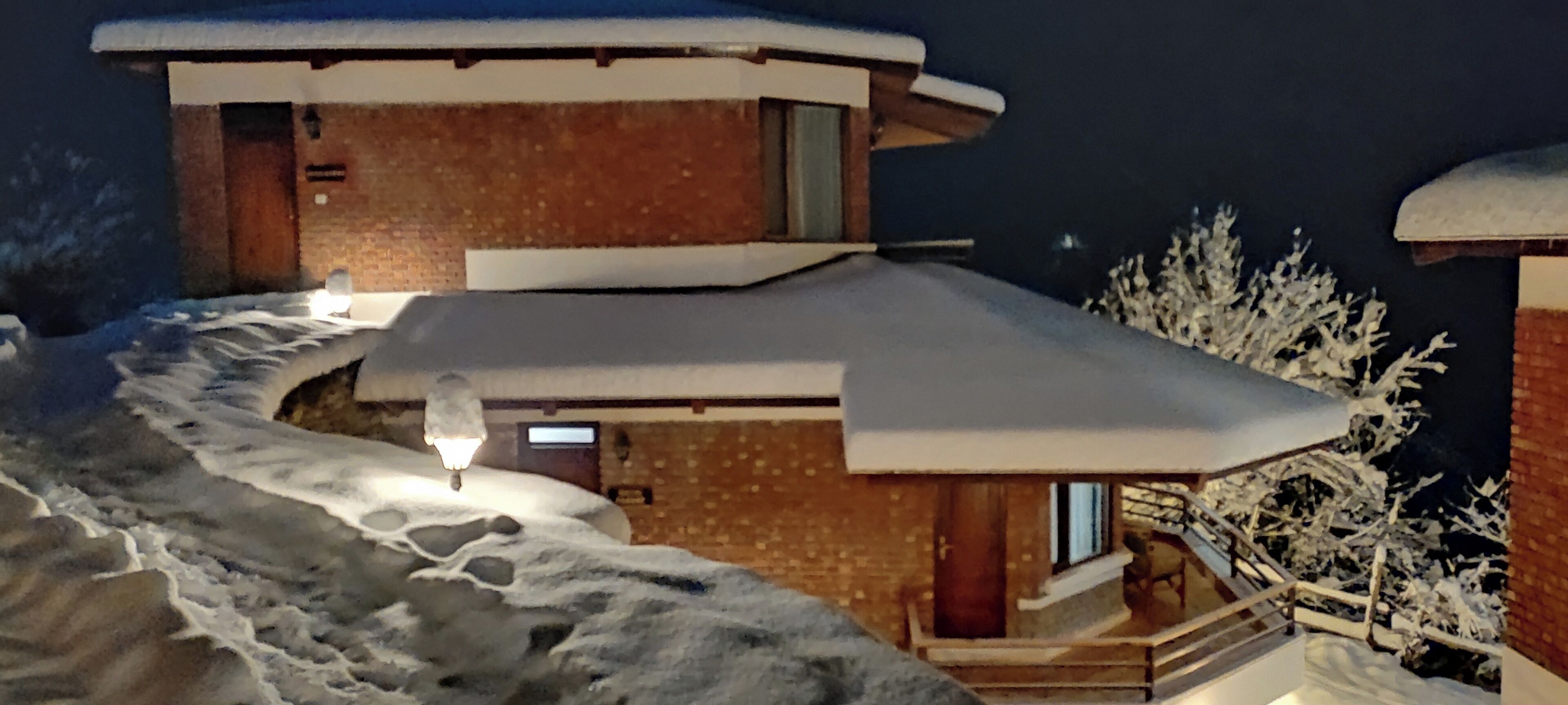 Edifício coberto de neve com fachada de madeira e telhado nevado. Uma escadaria leva à entrada, e uma luz é visível no interior.