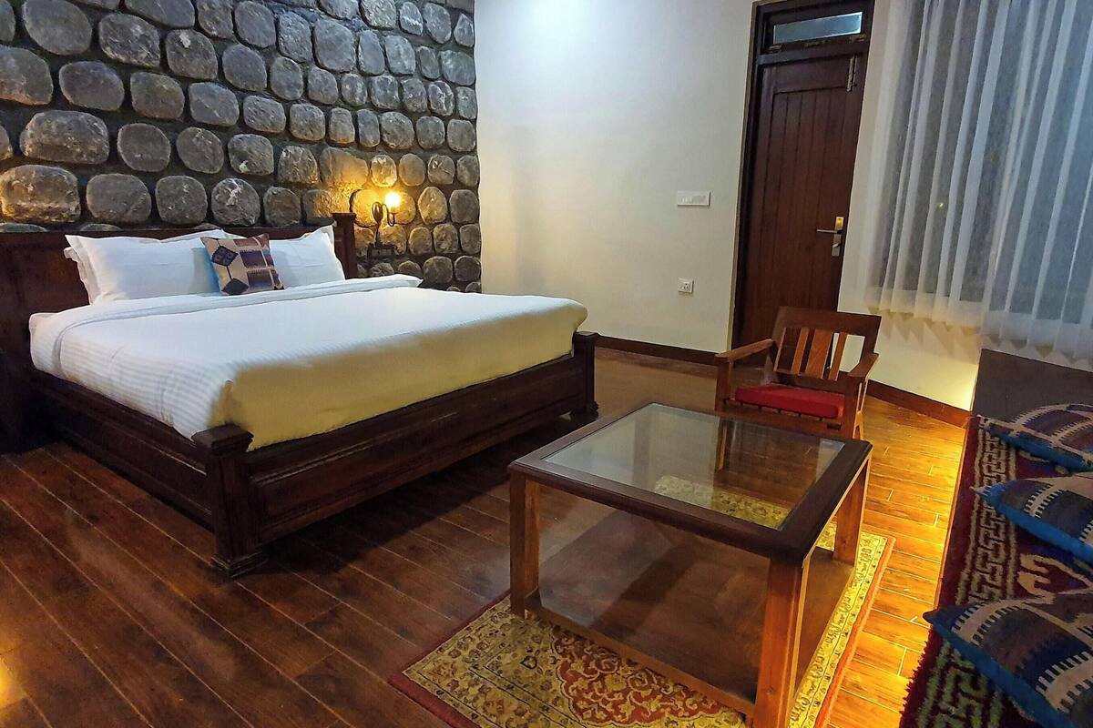 Una habitación con pared de piedra, cama con ropa de cama blanca, una silla de madera, una mesa de vidrio y una alfombra estampada.