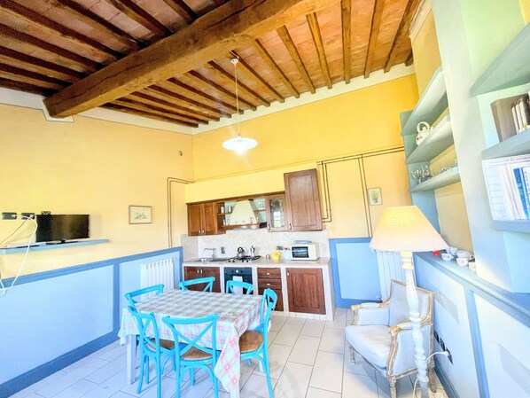 A compact kitchen with wooden cabinets, a dining table with chairs, a TV mounted on the wall, and a lamp on a side table. - Apt 5 Aircon, Wifi, Infinity Pool, Jacuzzi, Gardens Incredible! Sleeps 4 (Spoleto)