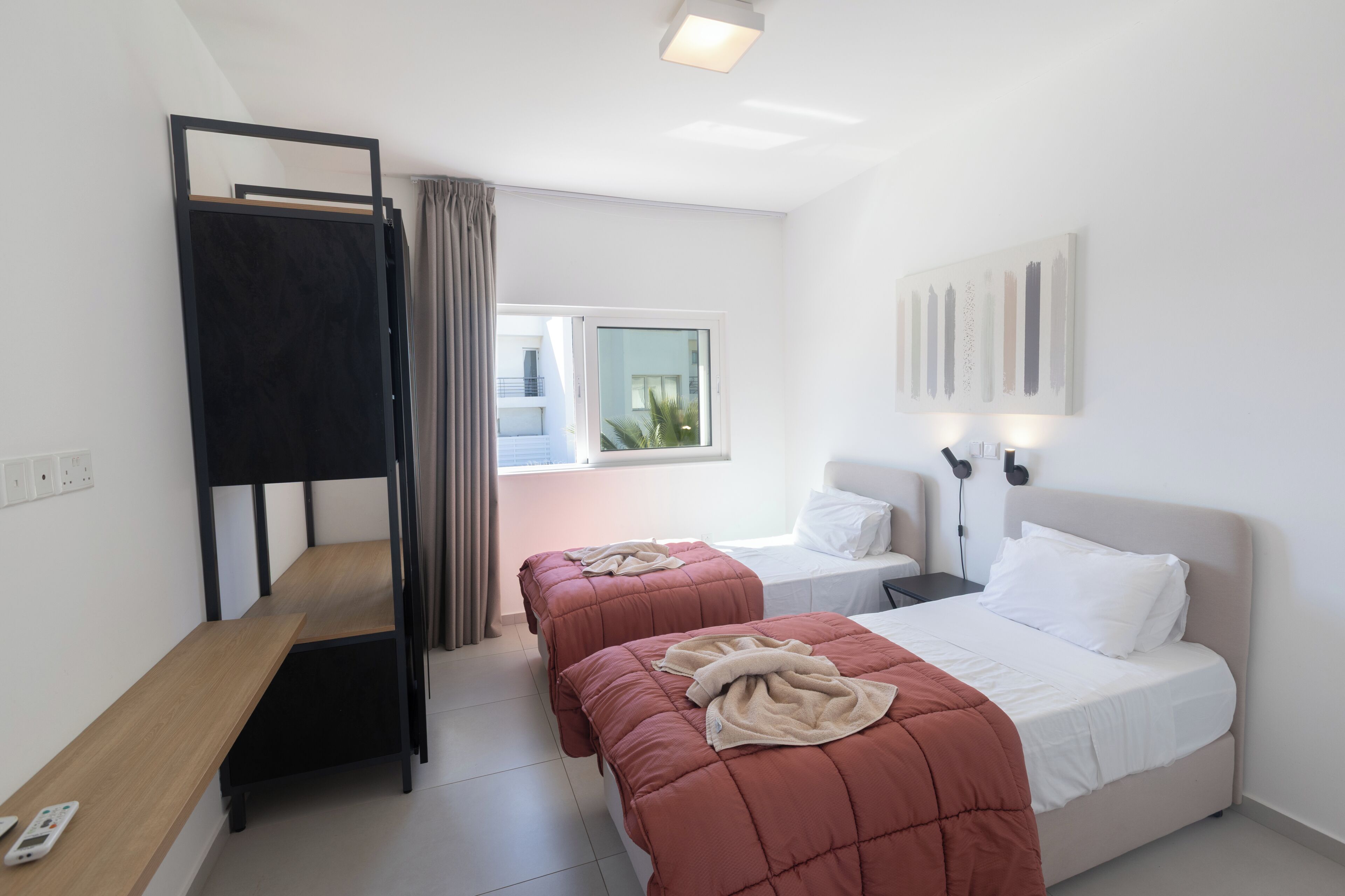 A bedroom with two beds, a bunk bed, a bedside table, a window, and a wall-mounted light fixture.