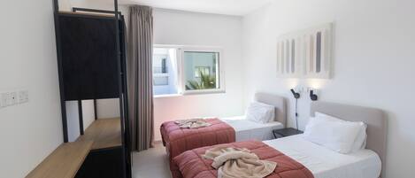 A bedroom with two beds, a bunk bed, a bedside table, a window, and a wall-mounted light fixture.