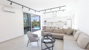 A modern living room with a sofa, armchair, and coffee table, a kitchen in the background, and a sliding glass door leading to an outdoor area.