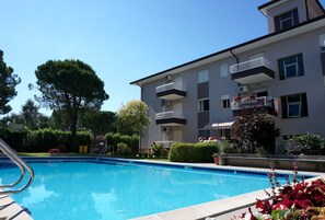 A swimming pool surrounded by a landscaped garden, with a multi-story building featuring balconies and air conditioning units.