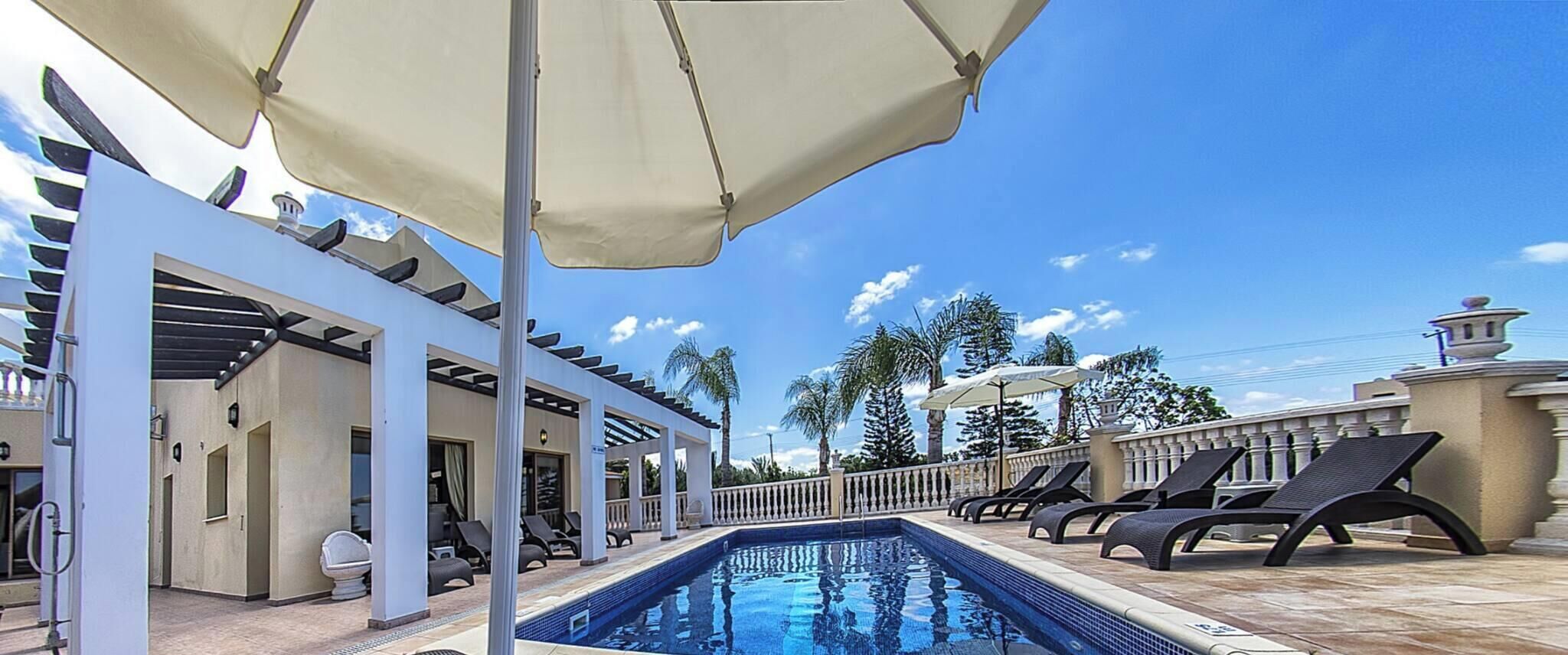 Zona de piscina con sillones de descanso y sombrillas, un paseo cubierto y un cielo azul despejado.