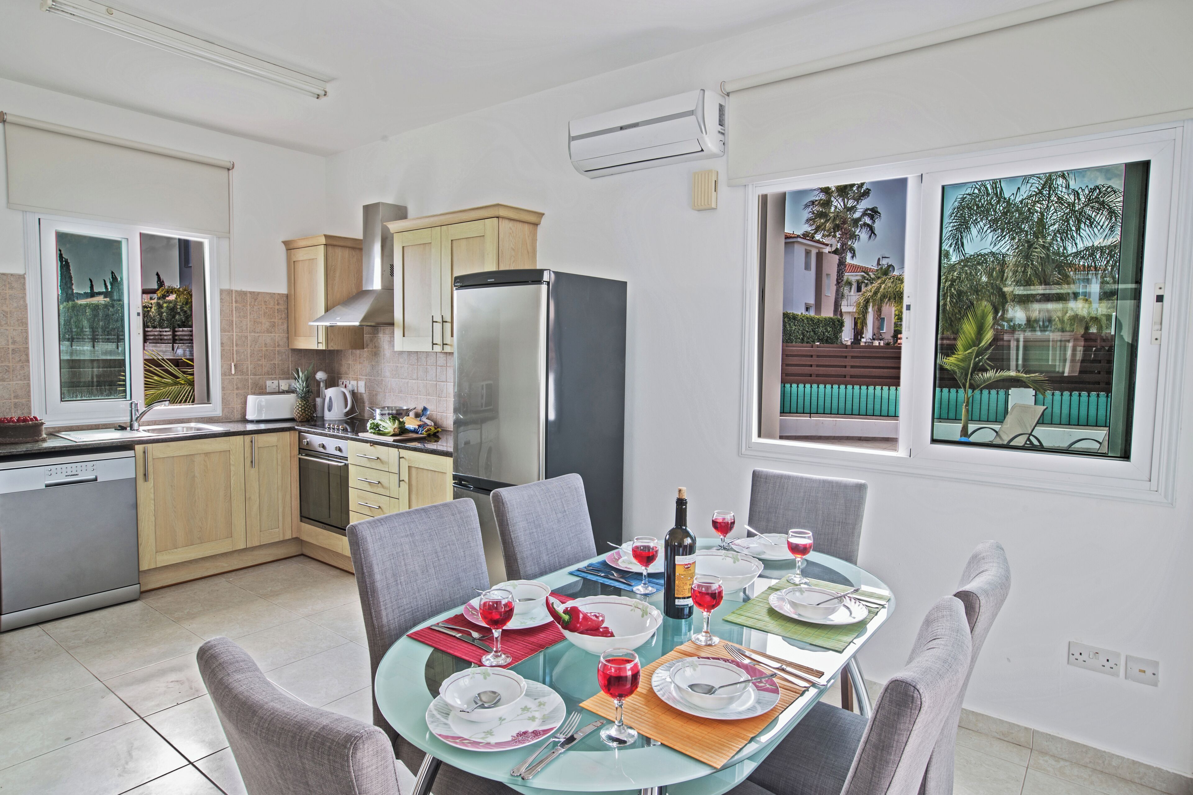 A modern kitchen with a dining area, featuring a glass table, grey chairs, and a view of a residential area.