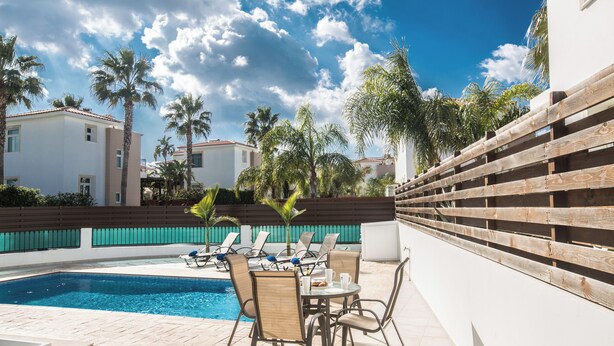 A pool area with lounge chairs and a table, surrounded by palm trees and a wooden fence. - Protaras Holiday Villa Me25 (Paralimni)