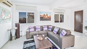 A modern living room with a sectional sofa, a coffee table, and decorative pillows. There are two windows with views of the outside, and a door to the right. - Protaras Holiday Villa Me25 (Paralimni)