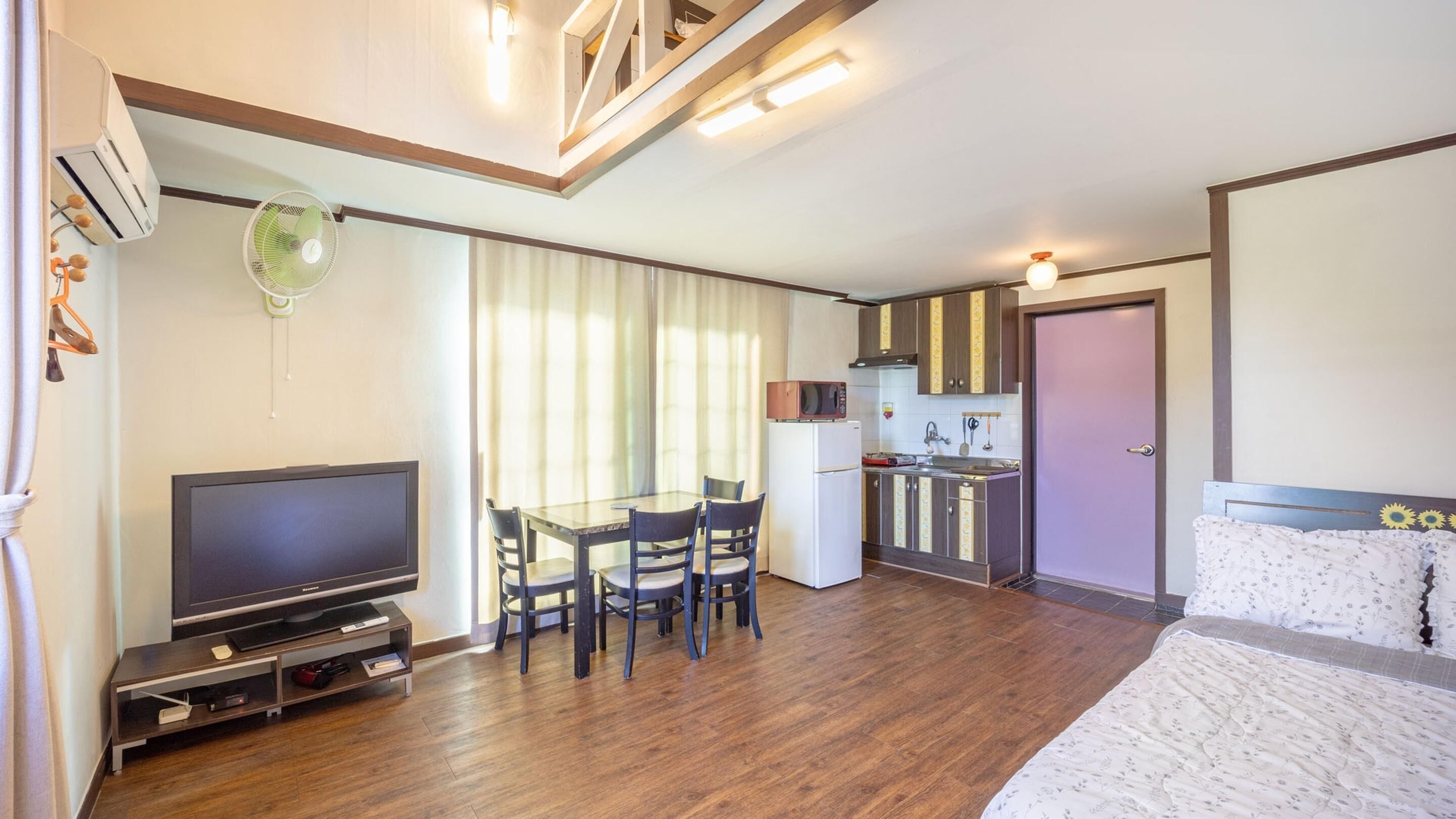 A compact room with a TV, dining table, chairs, a kitchenette, and a bed.