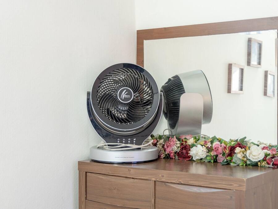 A standing fan on a wooden dresser with a mirror above it.