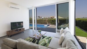 Ein modernes Wohnzimmer mit Flachbildfernseher, einer Couch und einem Esstisch mit Blick auf einen Swimmingpool und die umliegende Natur.