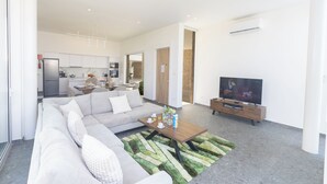Ein modernes Wohnzimmer mit einer Couch, einem Couchtisch und einem Fernseher. Im Hintergrund ist eine Küche mit Schränken und einem Kühlschrank zu sehen.