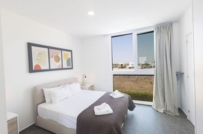 A modern hotel room with a large bed, a nightstand, a window with curtains, and a city view. - Protaras Vie Bleu Villa Vb15 (Paralimni)
