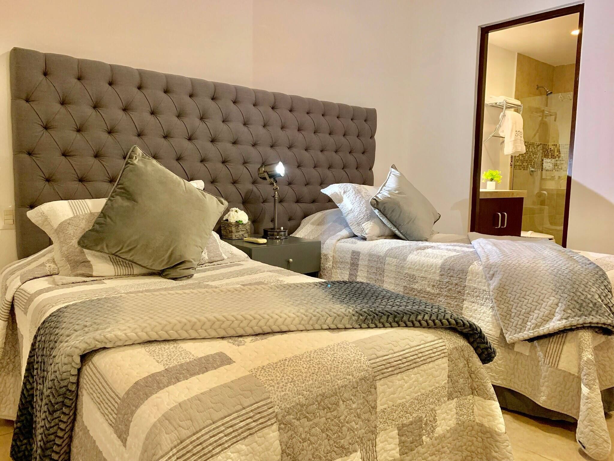 A bedroom with a tufted headboard, two beds with grey bedding, a bedside table with a lamp, and a mirror reflecting a bathroom.