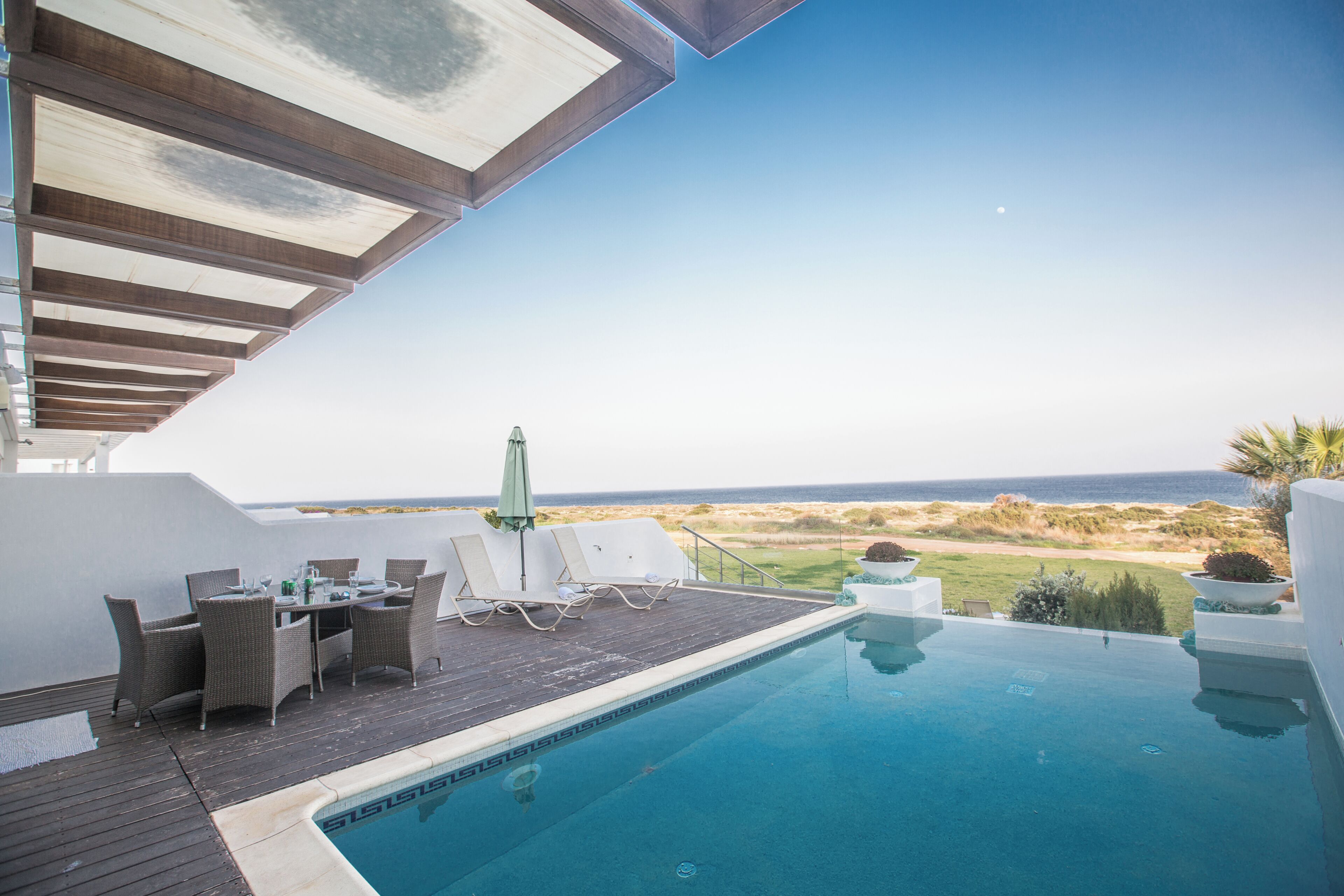 A pool with a view of the ocean, a covered patio with chairs and a table, and a grassy area.