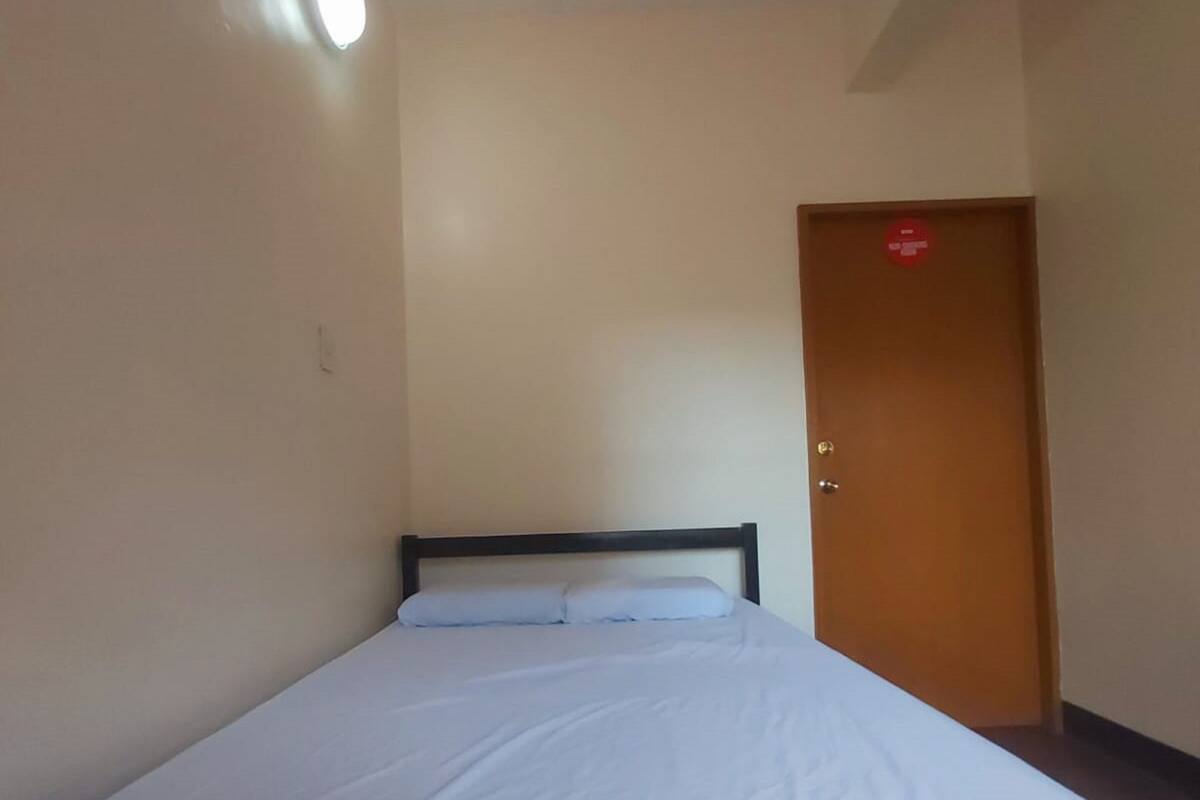 A single bed in a room with a wooden headboard, a door with a red sign, and a ceiling light.