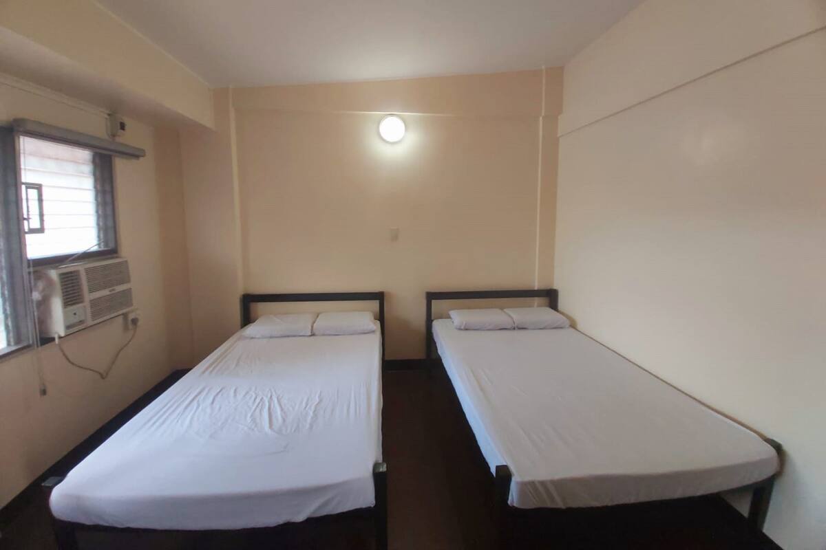 A room with two single beds, a window with an air conditioning unit, and a light fixture on the ceiling.