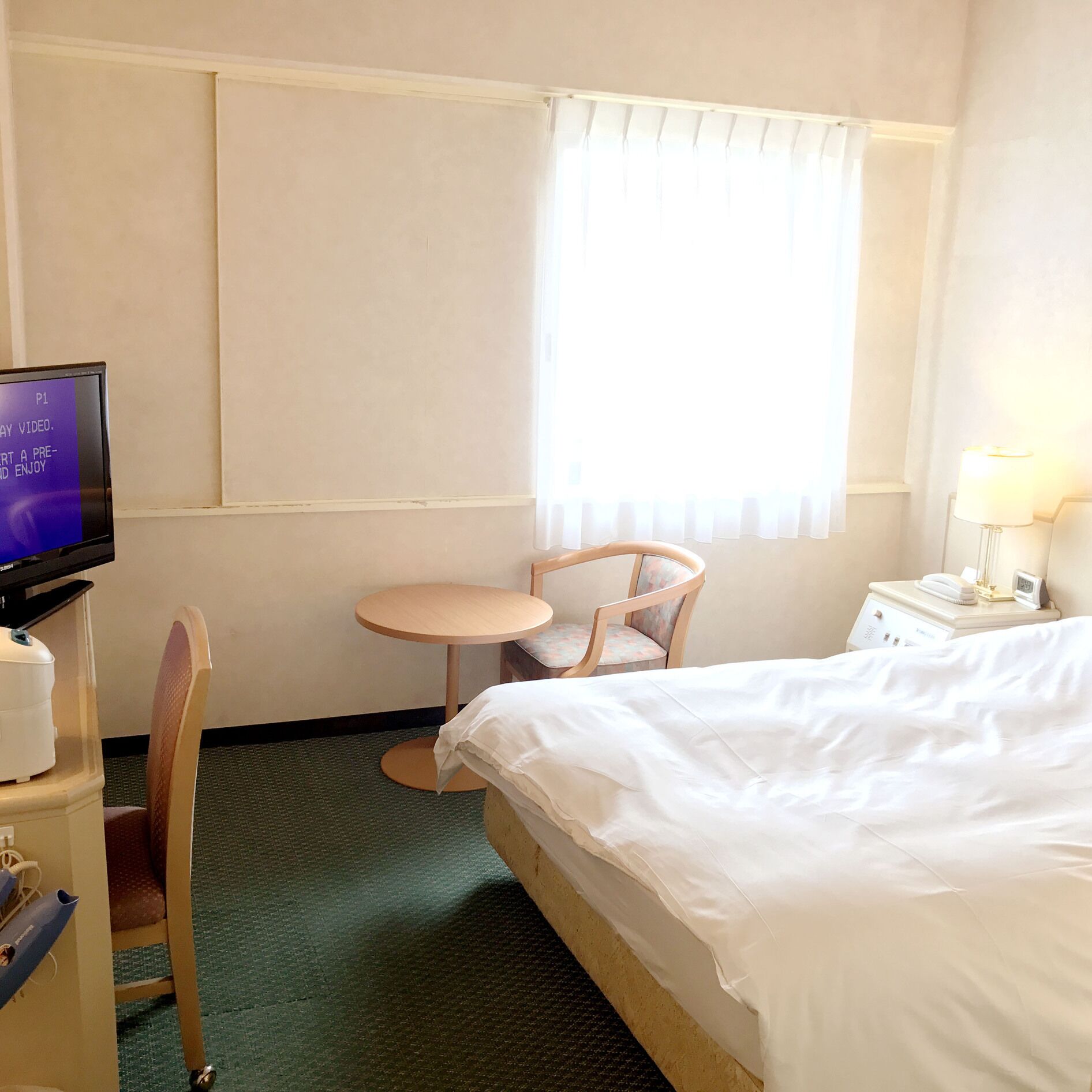 Ein Hotelzimmer mit einem Bett, einem kleinen runden Tisch, einem Sessel, einem Fernseher und einem Nachttisch mit Lampe.