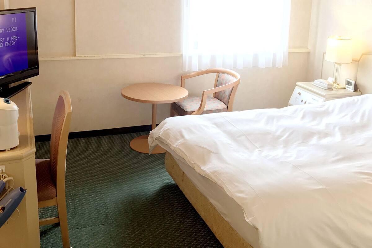 Hotellihuone, jossa on sänky, pieni pyöreä pöytä, tuoli, televisio ja yöpöytä, jossa on lamppu.