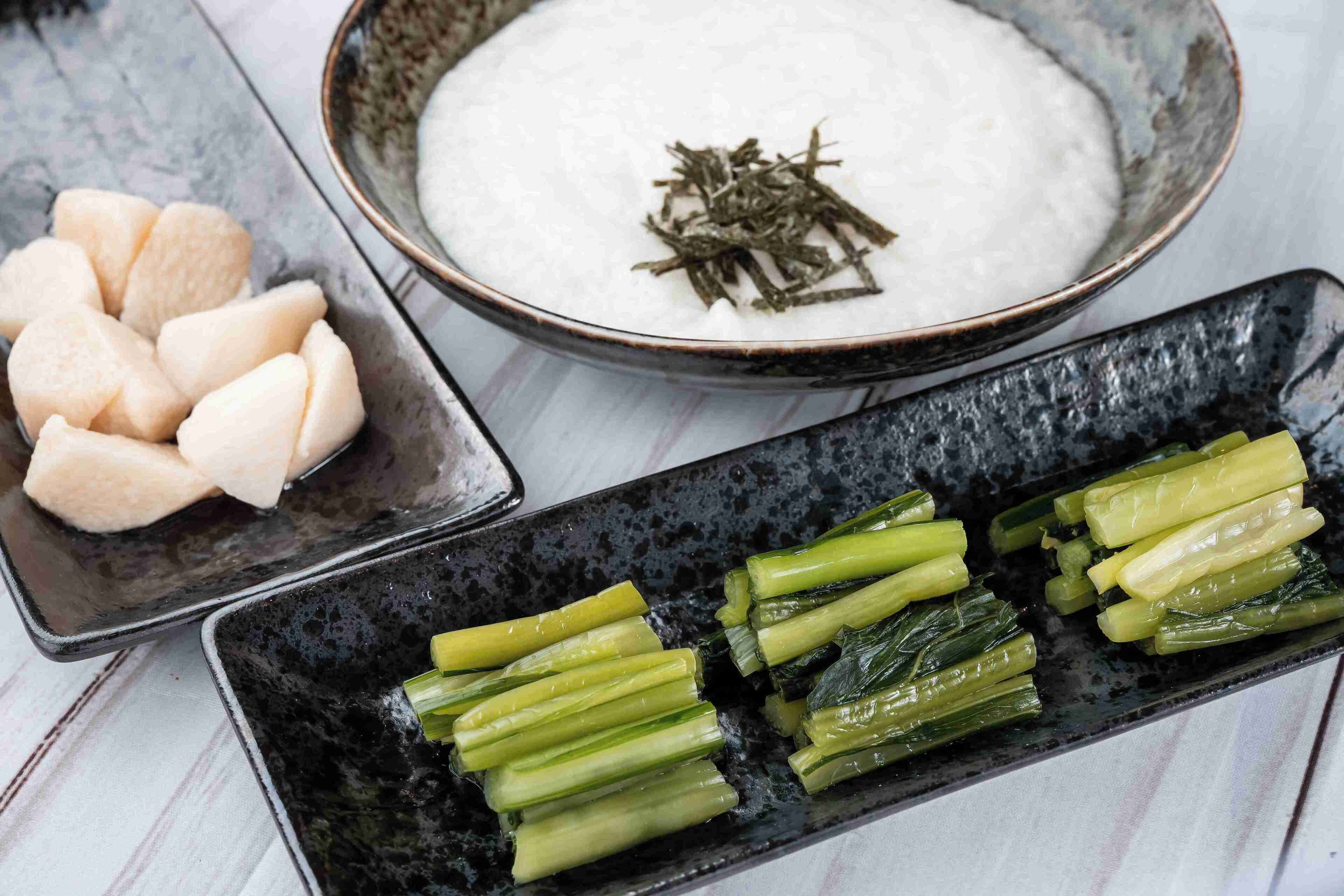 A bowl of white rice with seaweed garnish, a plate of sliced green vegetables, and a bowl of sliced white radish.